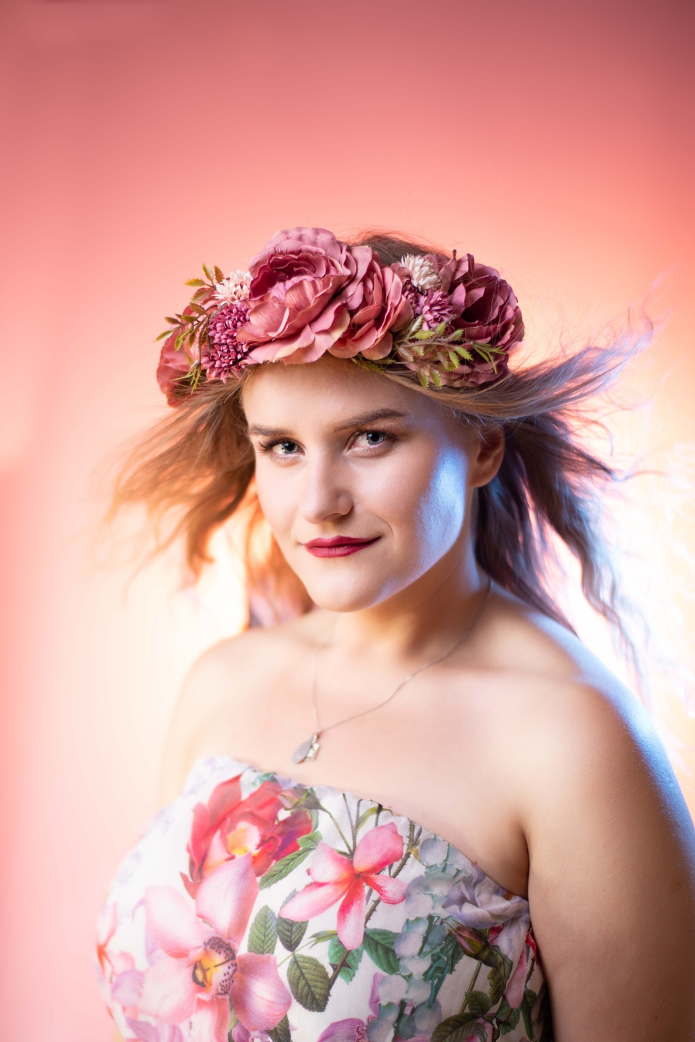 Portret kobiety z wiankiem z różowych kwiatów na głowie, w sukience w kwiaty, na rozmytym różowo-pomarańczowym tle, z wiatrem we włosach.