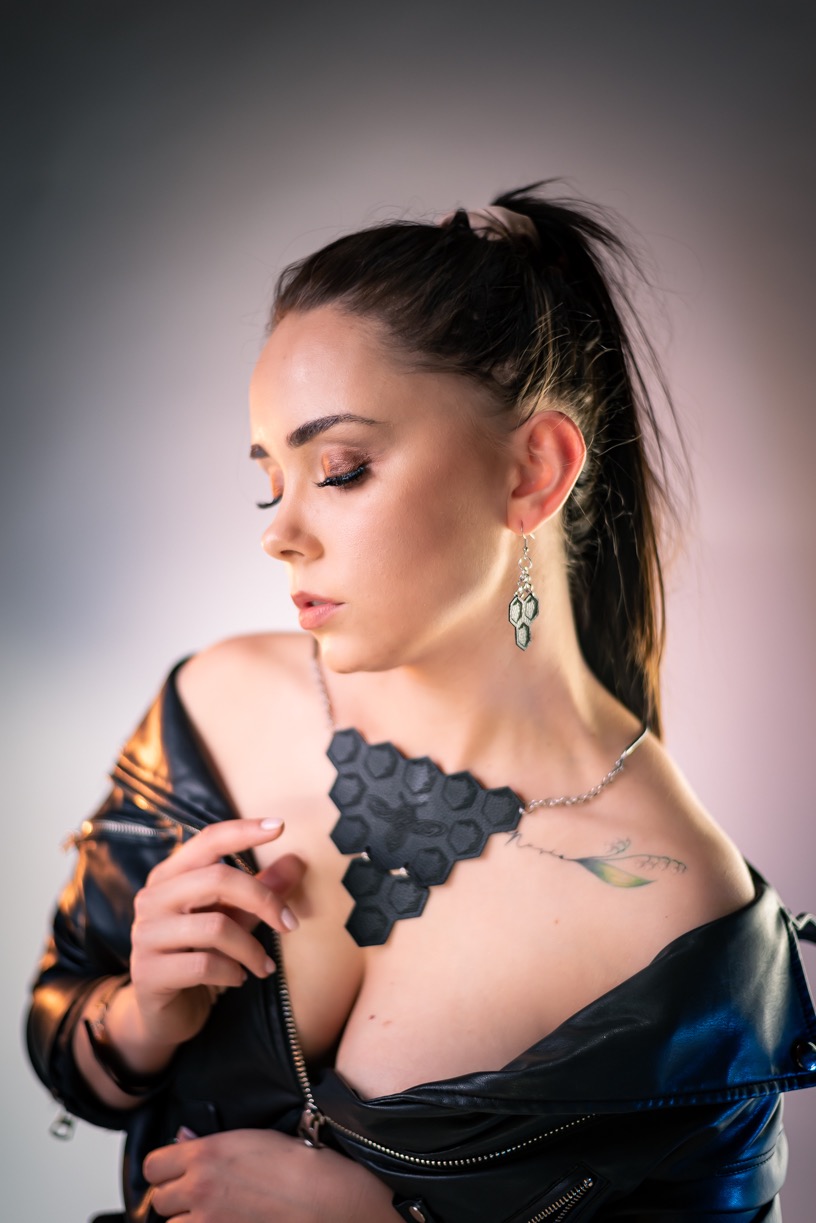 Portret kobiety z ciemnymi włosami w kucyku, ubranej w skórzaną kurtkę, z geometrycznym naszyjnikiem i kolczykami w kształcie plastra miodu, z delikatnym makijażem i tatuażem na dekolcie.
