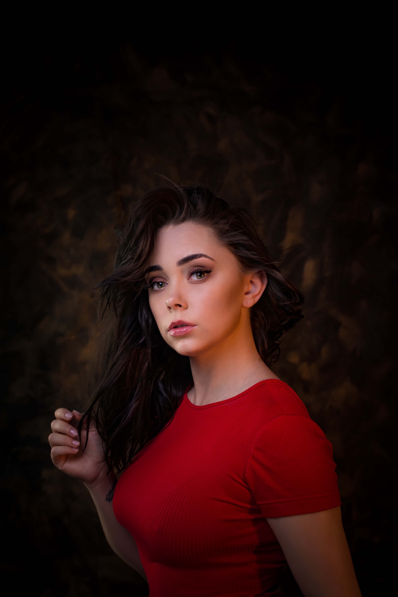 Portret młodej kobiety w czerwonej koszulce na ciemnym, rozmytym tle. Delikatny makijaż, rozwiane włosy i subtelny uśmiech.