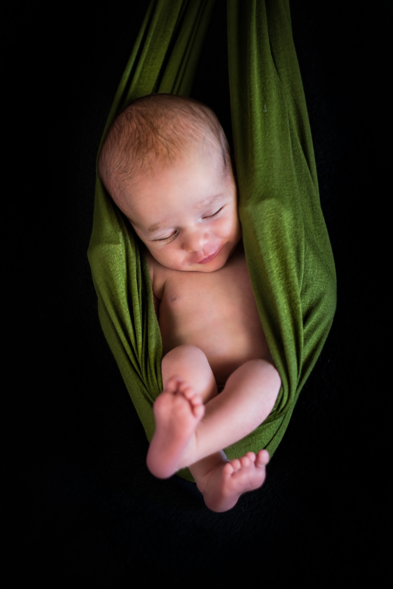 Śpiący noworodek zawinięty w zielony materiał na czarnym tle, delikatny uśmiech na twarzy dziecka.