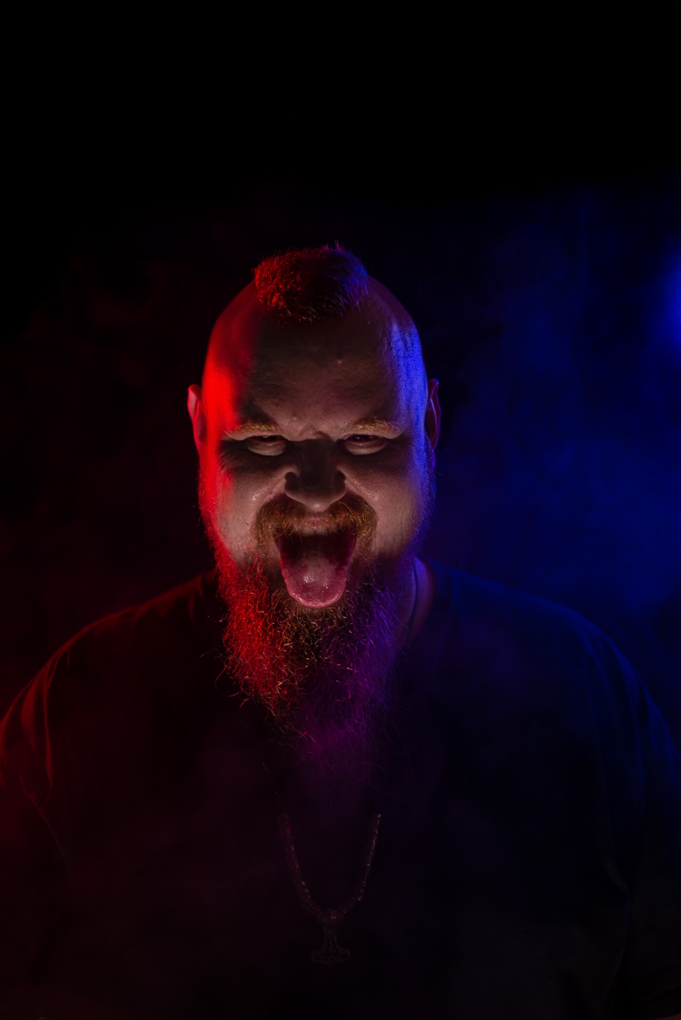 Portret mężczyzny z czerwoną brodą i irokezem, wystawiającego język, oświetlonego czerwonym i niebieskim światłem.