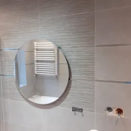 Jasna łazienka w trakcie wykańczania, widoczne płytki w odcieniach beżu, okrągłe lustro odbijające grzejnik drabinkowy, niezabezpieczone otwory instalacyjne w ścianie.