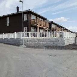 Szeregowe, dwukondygnacyjne domy z ciemnego drewna z balkonami i białym ogrodzeniem na kamiennej podmurówce, widziane z ulicy w słoneczny dzień.