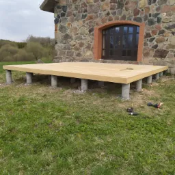 Surowa drewniana konstrukcja tarasu na betonowych podporach, narzędzia budowlane na trawie, kamienny budynek w tle.