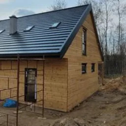 Surowy, drewniany dom w trakcie budowy, z metalowym dachem, widoczne rusztowania i teren budowy.