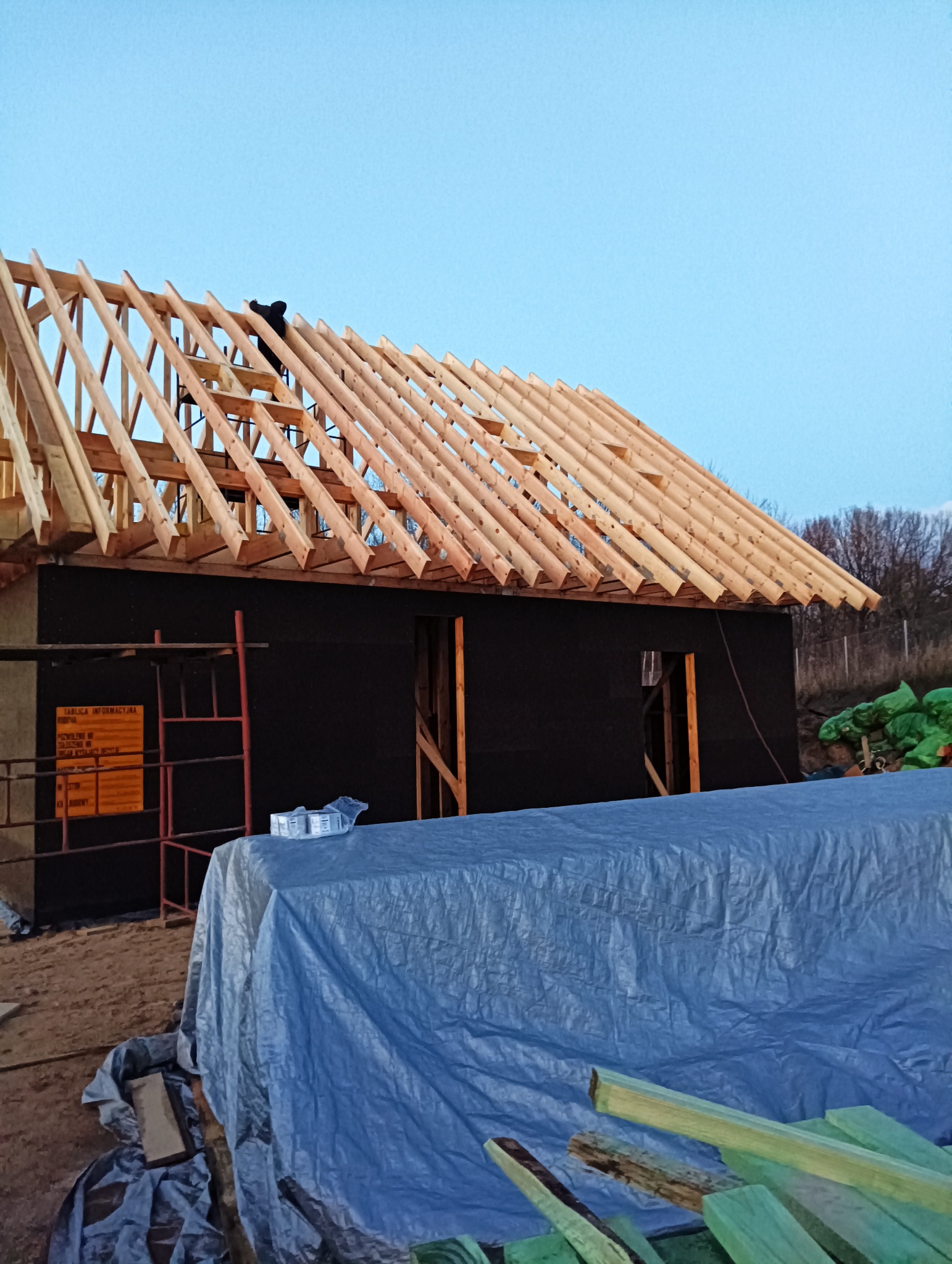 Wznoszenie drewnianej konstrukcji dachu na domu w trakcie budowy, widoczny pracownik na dachu, zabezpieczone materiały budowlane i rusztowanie.