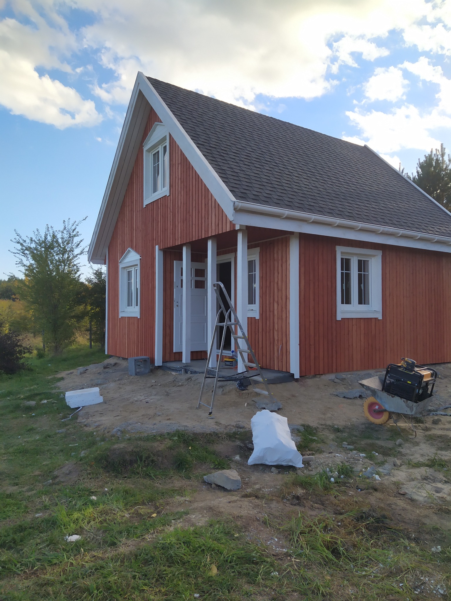 Nowo wybudowany, pomalowany na czerwono budynek z drewna, z białymi ramami okiennymi i drzwiami, na tle błękitnego nieba z chmurami, widoczne narzędzia budowlane i materiały budowlane.