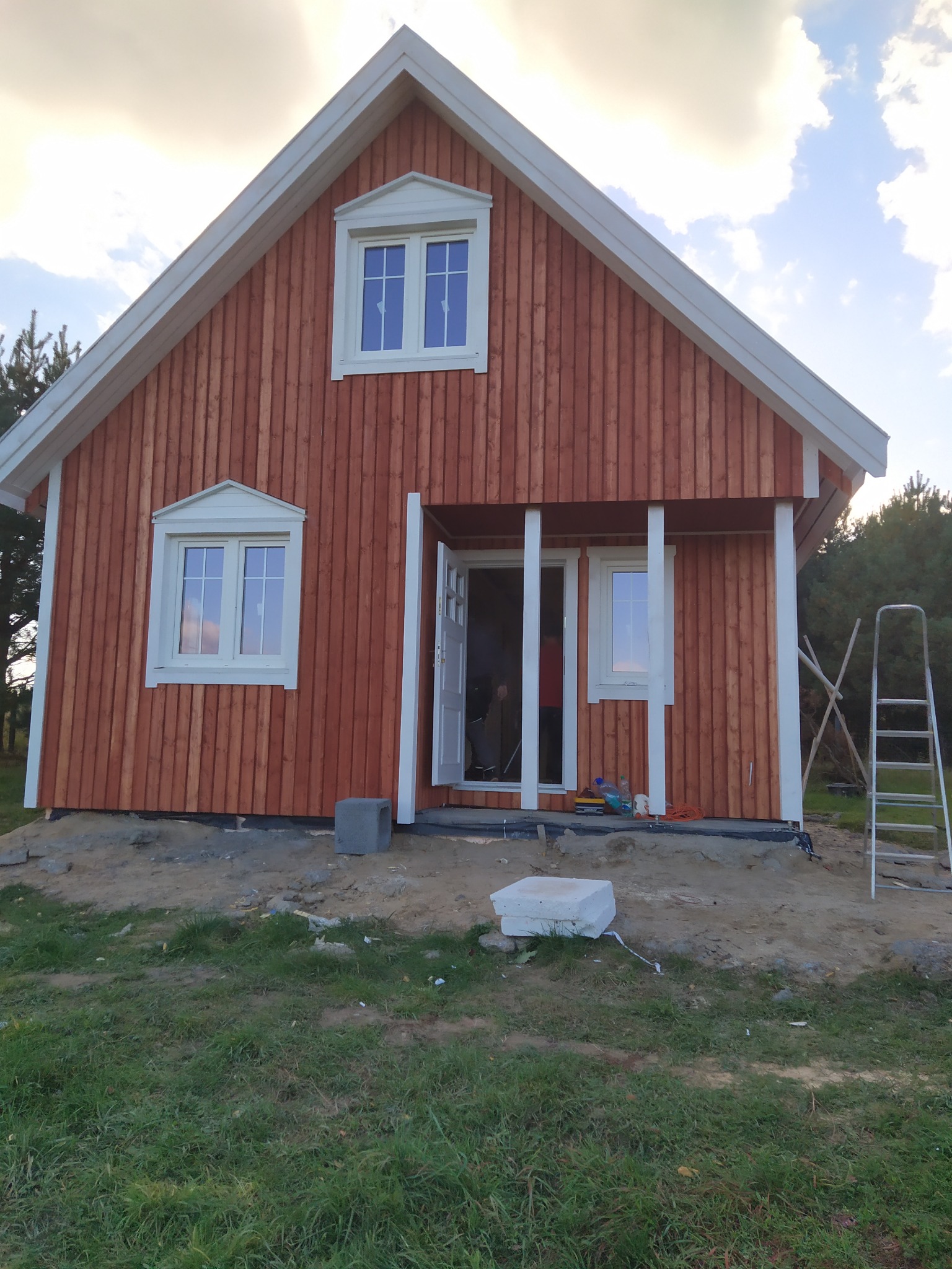 Surowy stan nowo wybudowanego domu z drewnianą elewacją w odcieniach brązu, białe obramowania okien i dachu, widoczne rusztowanie i teren w trakcie zagospodarowywania.