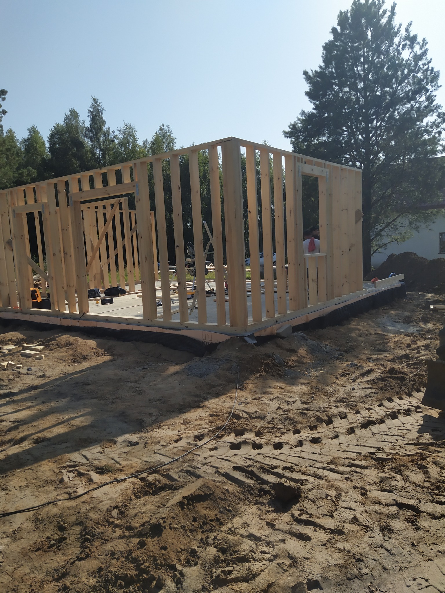 Wznoszenie konstrukcji domu z drewnianych belek na fundamencie, widoczne narzędzia i elementy konstrukcyjne, w tle drzewa i fragment budynku.