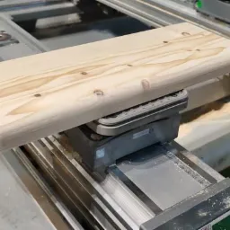 Obrabiany element z jasnego drewna o zaokrąglonych krawędziach, umieszczony na stole frezarki CNC z widocznymi śladami obróbki.