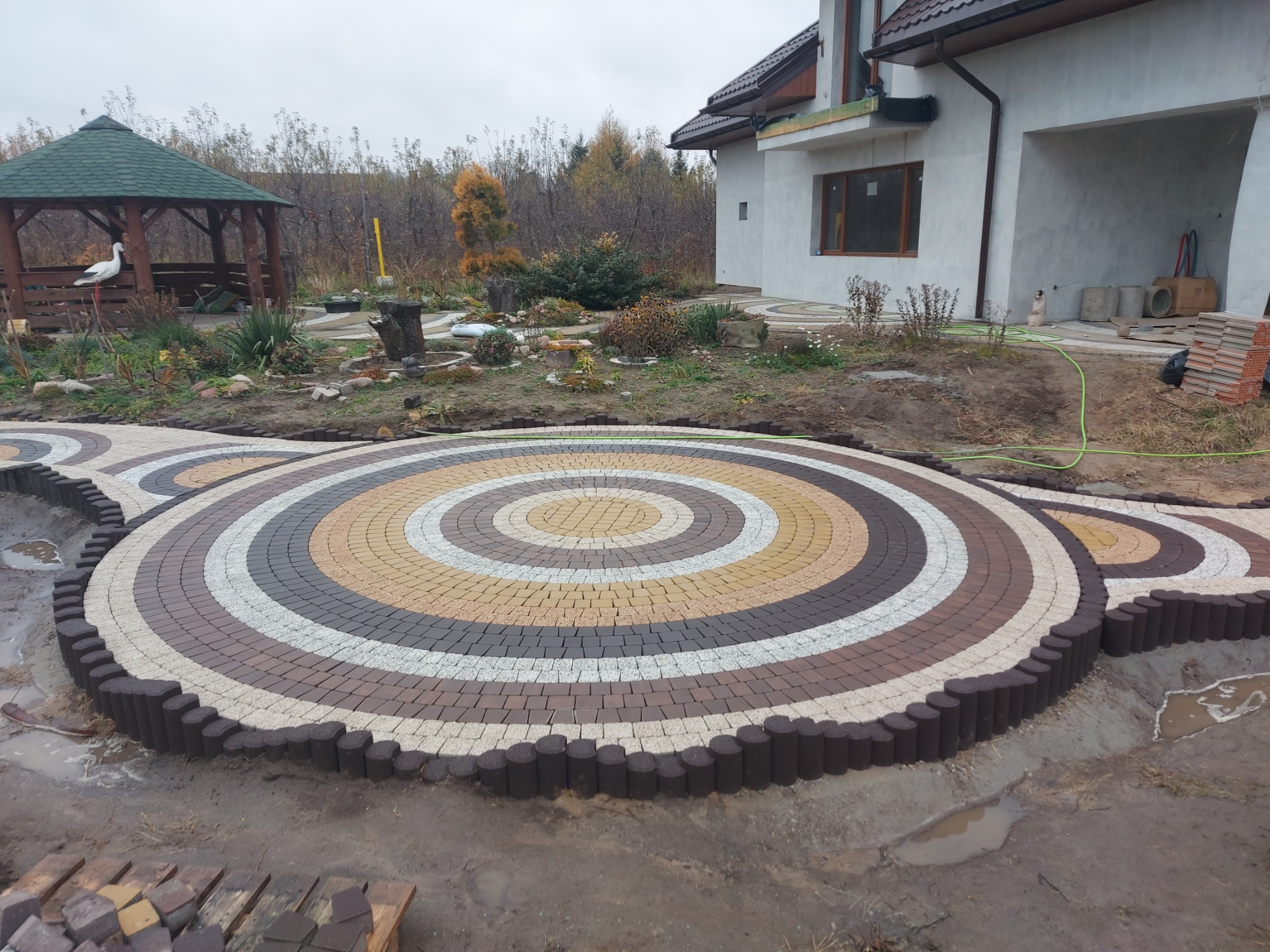 Okrągła kompozycja z kostki brukowej w odcieniach brązu, beżu i szarości, otoczona palisadą, na tle ogrodu z altaną i budynkiem w trakcie budowy.