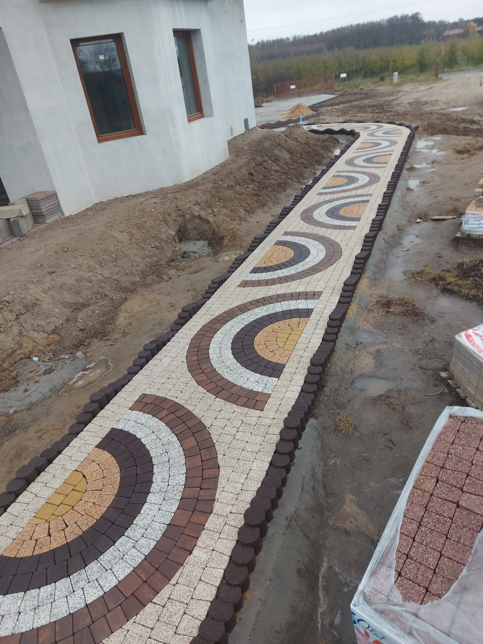 Ścieżka z kostki brukowej ułożona we wzór półkola, obrzeża z okrągłych kostek, widoczne palety z kostką, dom w tle, teren w trakcie prac ziemnych.