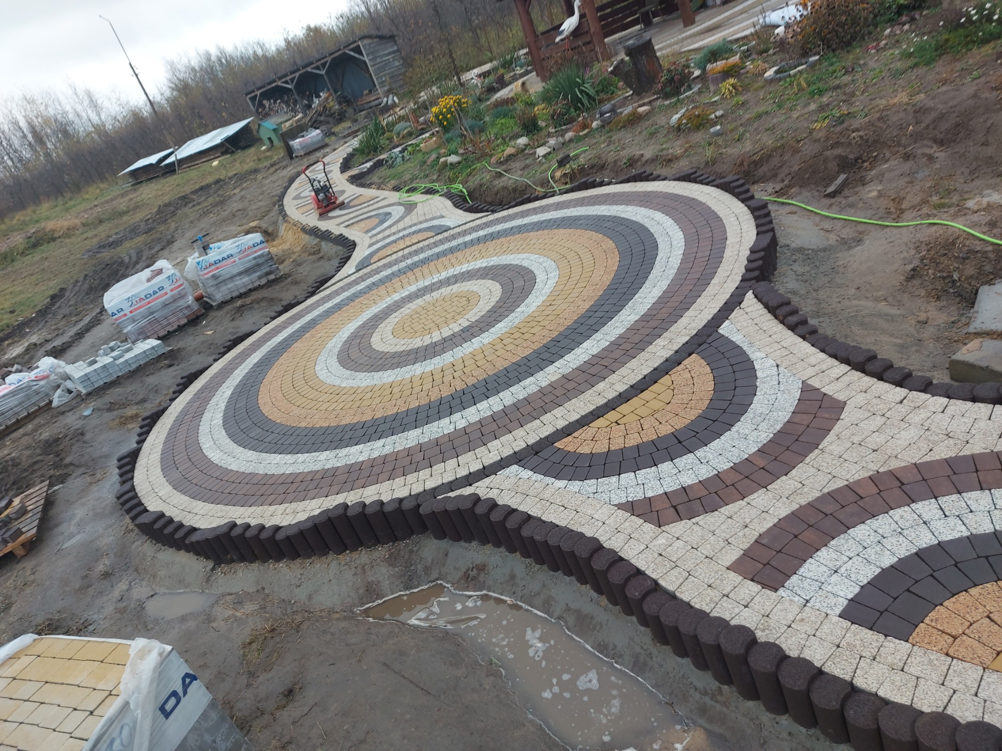 Okrągła ścieżka z kostki brukowej ułożona w spiralny wzór z brązowym obrzeżem, w tle trawnik, drzewa i maszyna do ubijania ziemi.