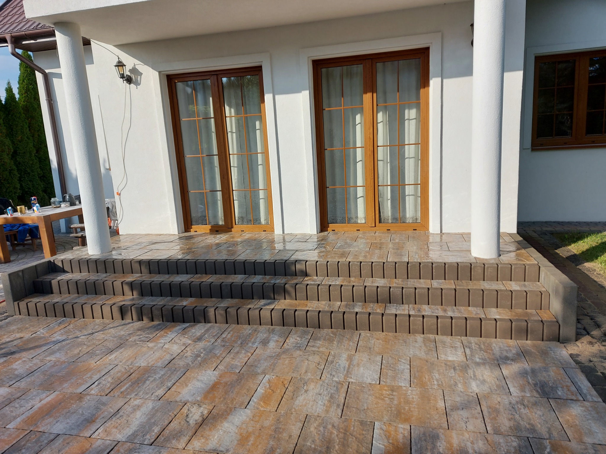 Ułożona kostka brukowa w odcieniach brązu i szarości prowadzi do domu z białymi kolumnami w Chynowie, tworząc schody wejściowe.