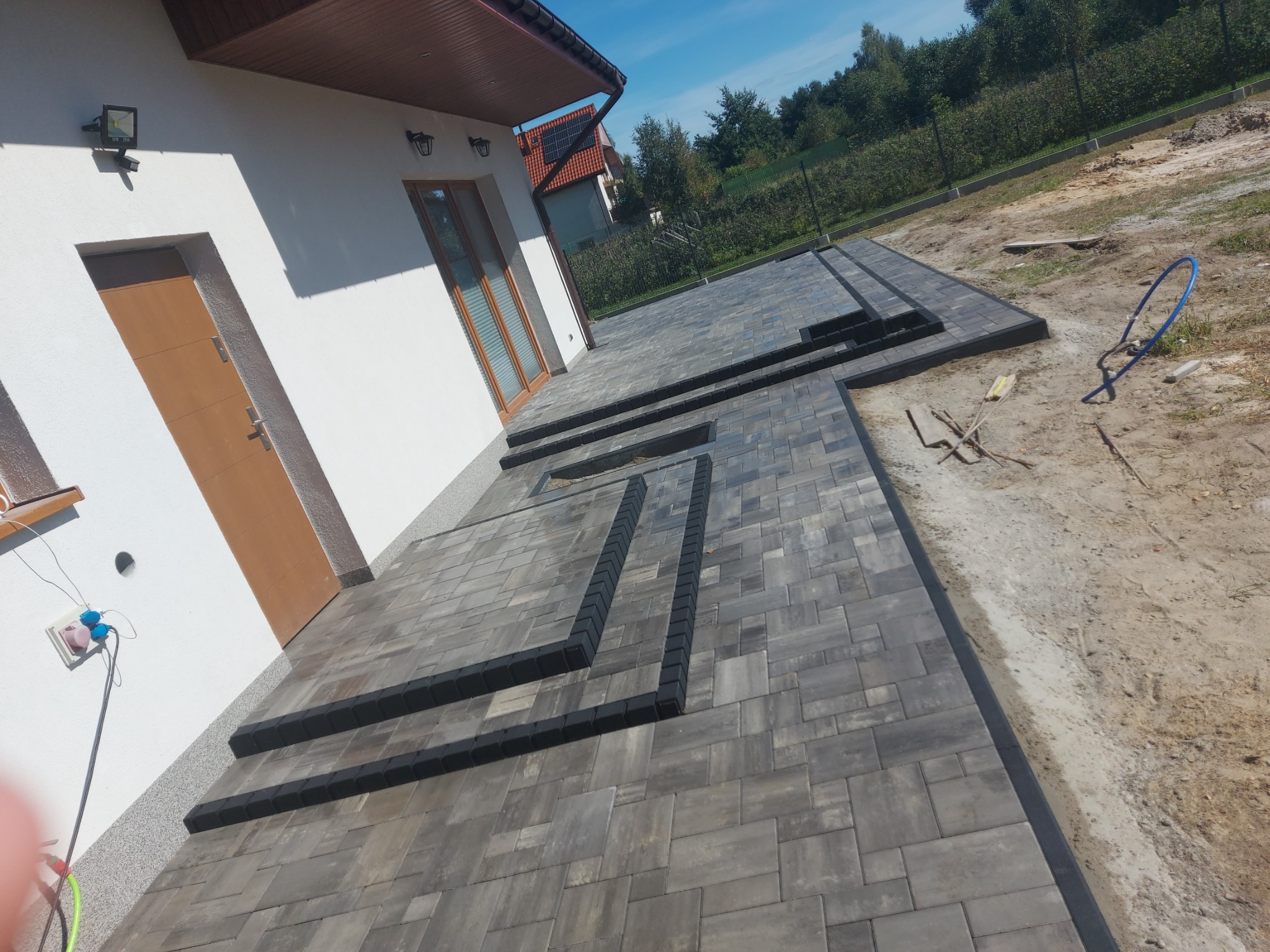 Świeżo ułożony taras z szarej kostki brukowej z czarnymi obrzeżami, widok z perspektywy domu, z widocznymi elementami otoczenia w trakcie prac wykończeniowych.