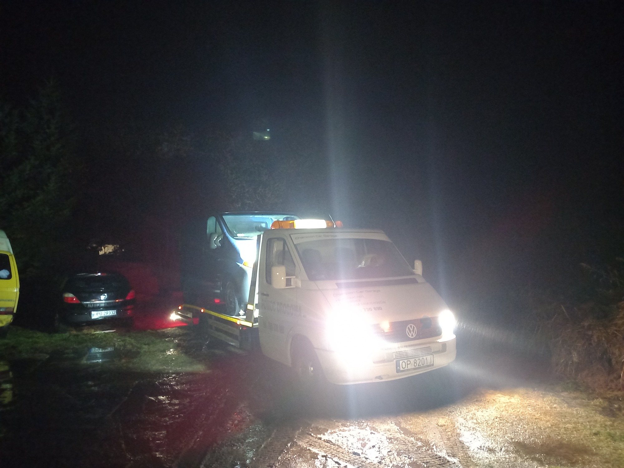 Samochód osobowy transportowany na lawecie pomocy drogowej w nocy, oświetlony reflektorami, na mokrej nawierzchni.