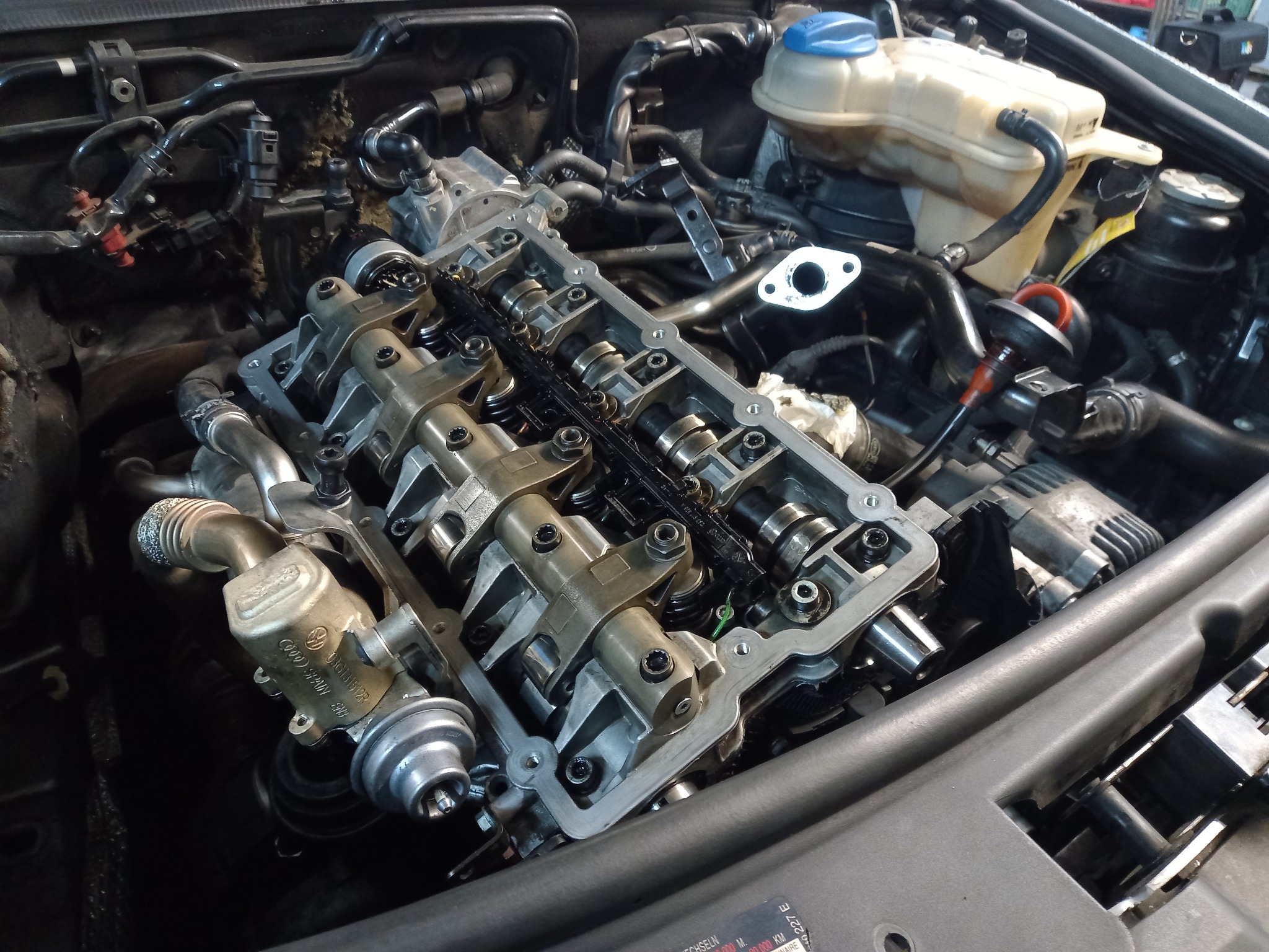 Widok z góry na otwarty silnik samochodowy z widoczną głowicą cylindrów i wałkami rozrządu, pokrytymi olejem, podczas naprawy w warsztacie.