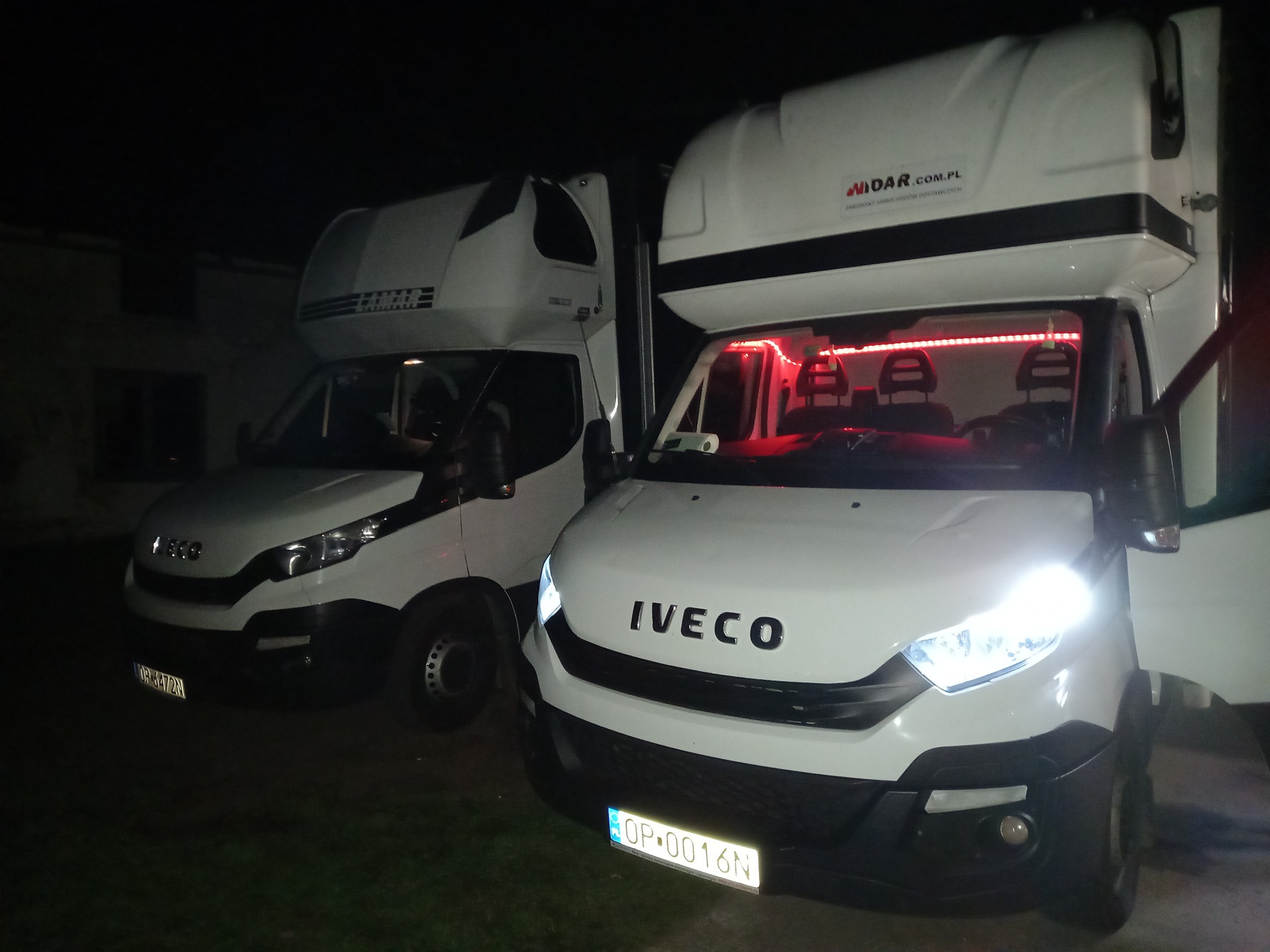 Dwa białe samochody dostawcze marki Iveco stojące obok siebie w nocy, z widocznymi tablicami rejestracyjnymi i oświetleniem LED w kabinie jednego z pojazdów.