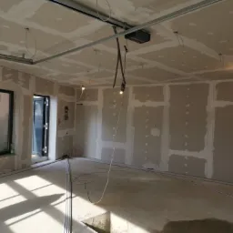 Wnętrze w trakcie montażu płyt kartonowo-gipsowych na ścianach i suficie, widoczne metalowe profile, szpachlowane łączenia płyt oraz wiszące kable oświetleniowe.