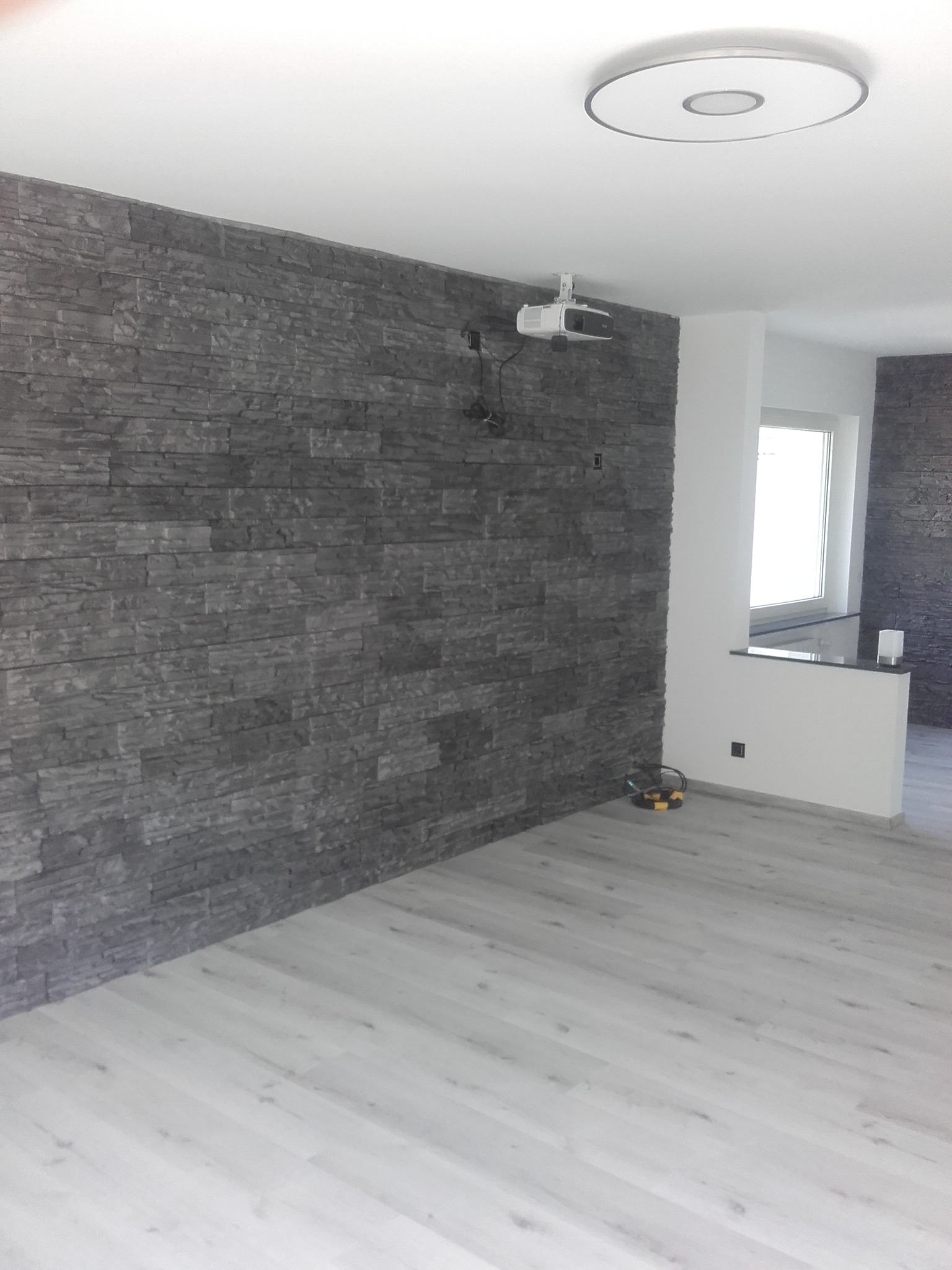 Szara ściana wykończona kamieniem dekoracyjnym, jasna podłoga, sufit z lampą i zamontowany projektor, widok na fragment pomieszczenia z oknem.