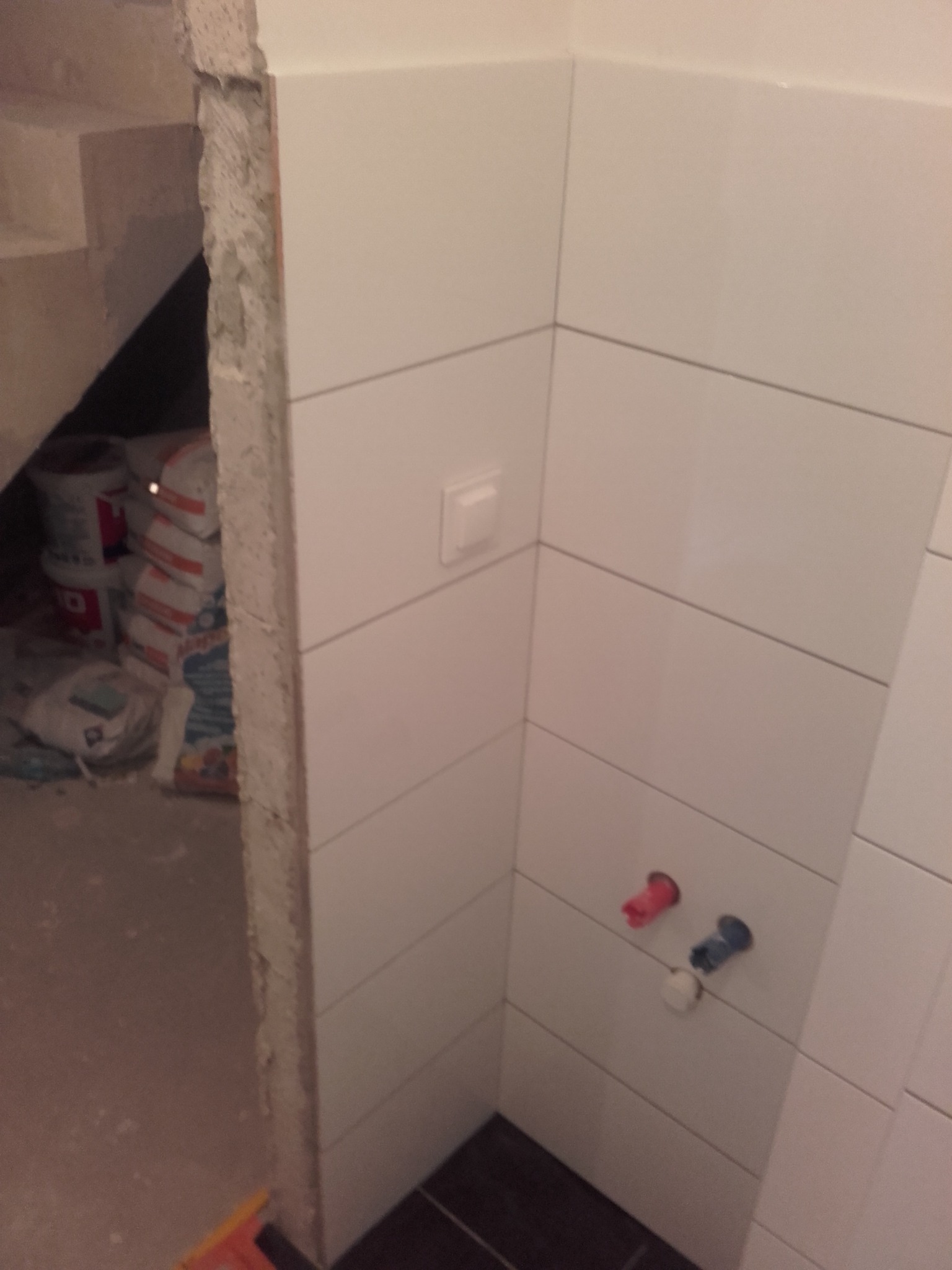 Białe, prostokątne płytki ścienne w trakcie układania w rogu łazienki, widoczne wystające rury z zaślepkami, w tle worki z zaprawą murarską i fragment surowego muru.