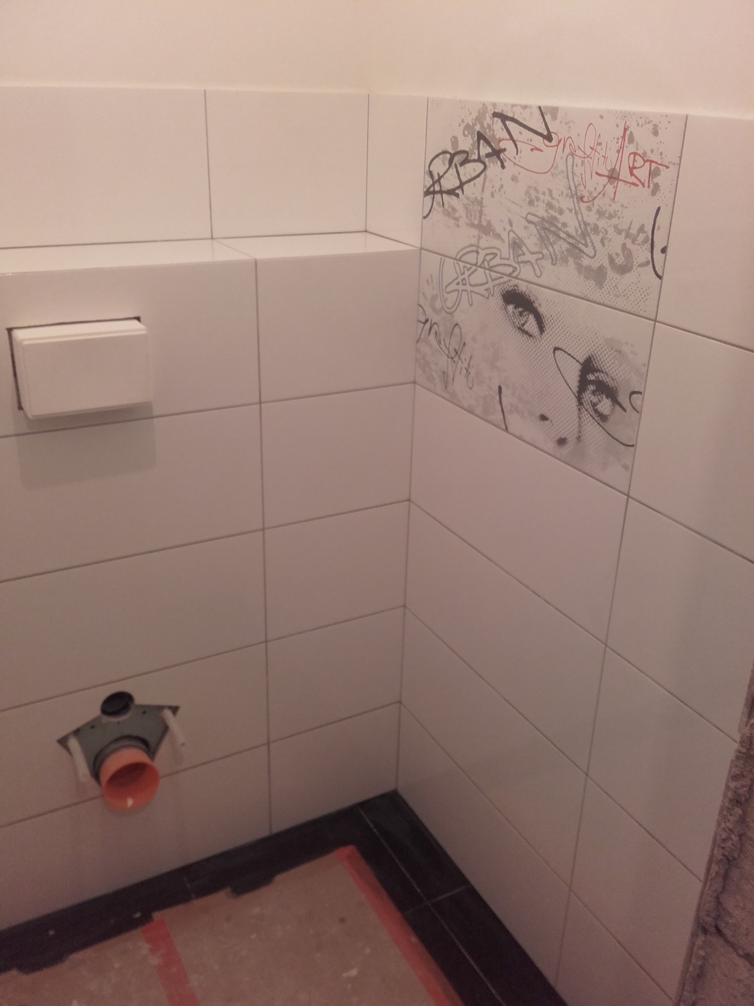 Fragment ściany w łazience z białymi, błyszczącymi płytkami ceramicznymi, dekoracyjna płytka z motywem graffiti i kobiecych oczu, widoczny częściowo zamontowany stelaż podtynkowy WC, brakująca...