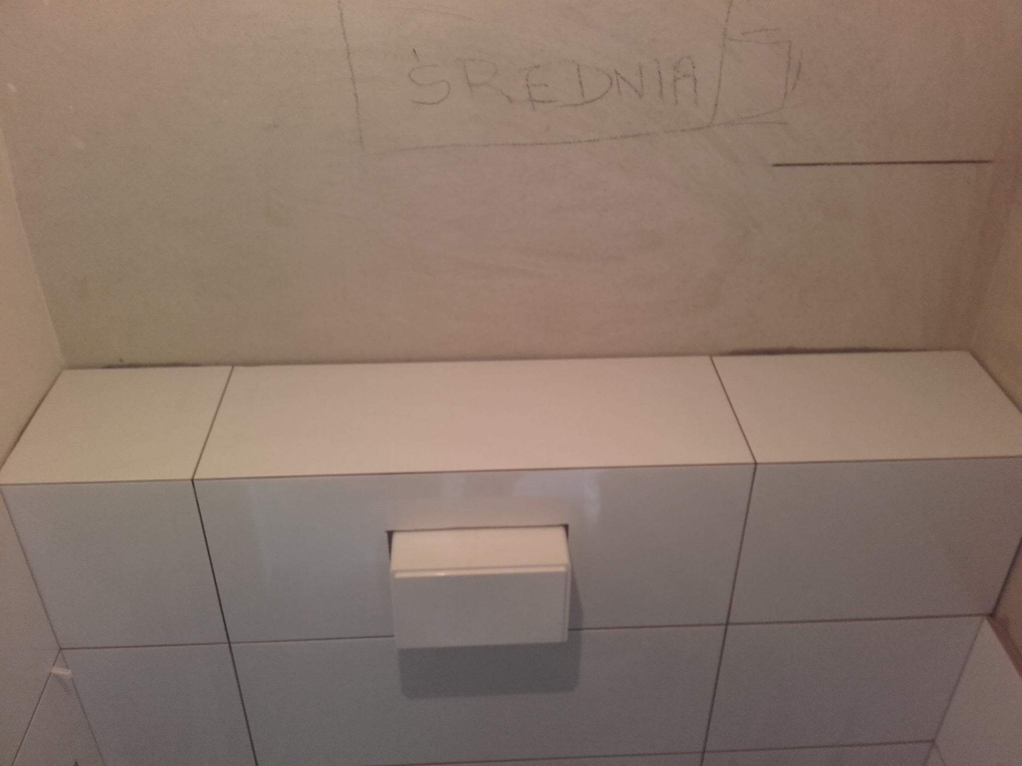 Świeżo ułożone, białe, błyszczące płytki ceramiczne na ścianie łazienki z zamontowanym uchwytem na papier toaletowy. Na górze ściany widoczny napis ołówkiem 'ŚREDNIA'.