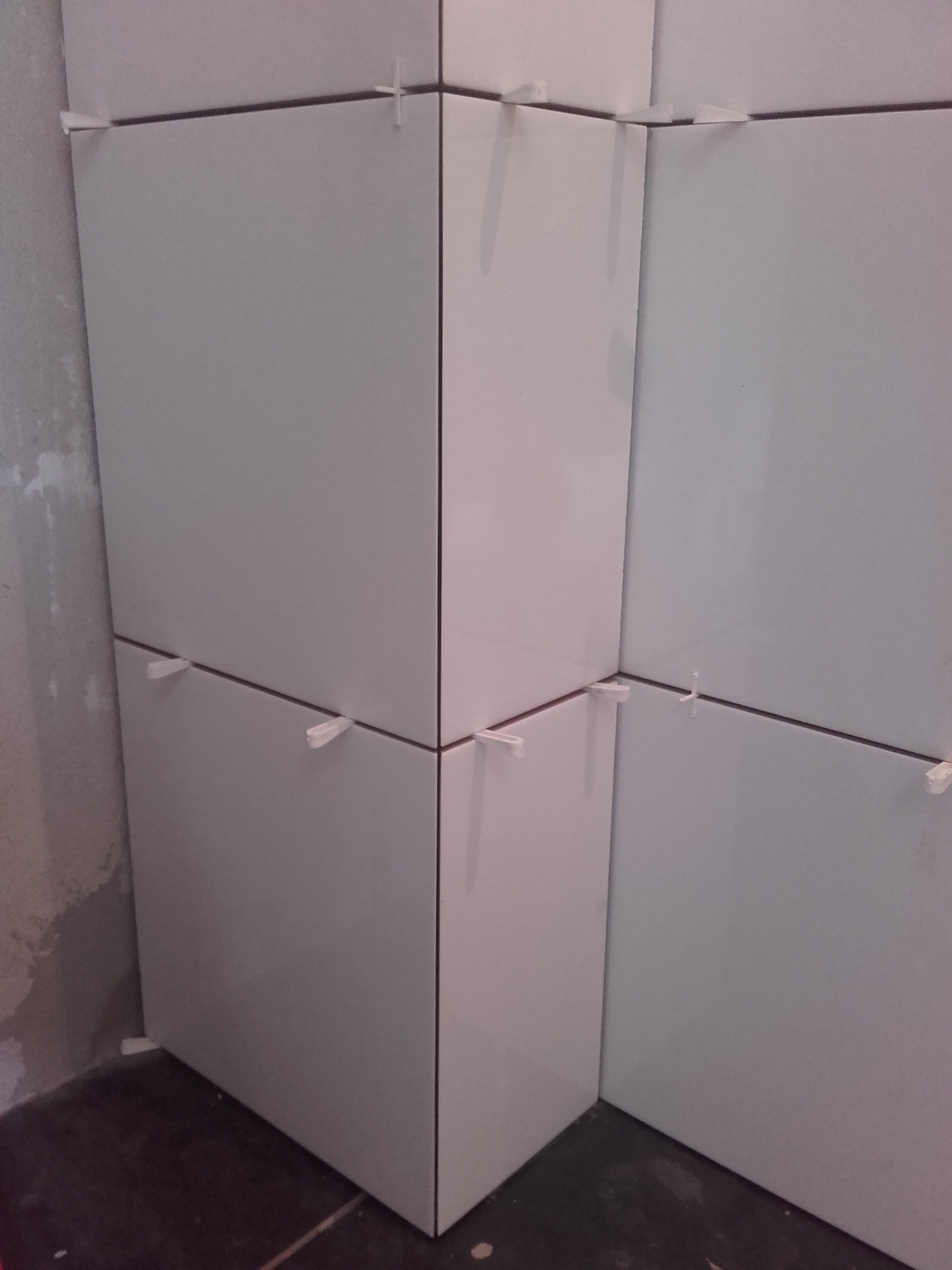 Fragment ściany w trakcie układania jasnych, kwadratowych płytek z widocznymi krzyżykami dystansowymi, dolna krawędź płytek styka się z ciemną podłogą, ściana obok surowa.