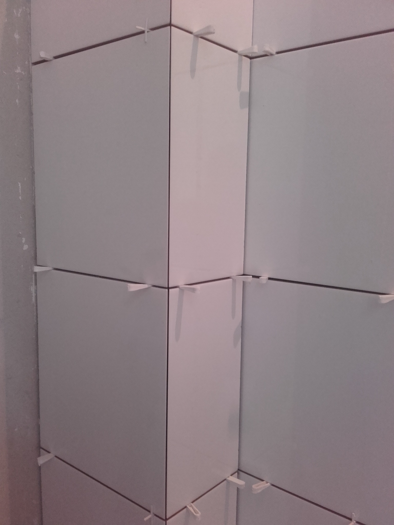 Świeżo ułożone, białe, kwadratowe płytki w rogu pomieszczenia, z widocznymi krzyżykami dystansowymi i ciemną fugą, na tle surowej ściany.