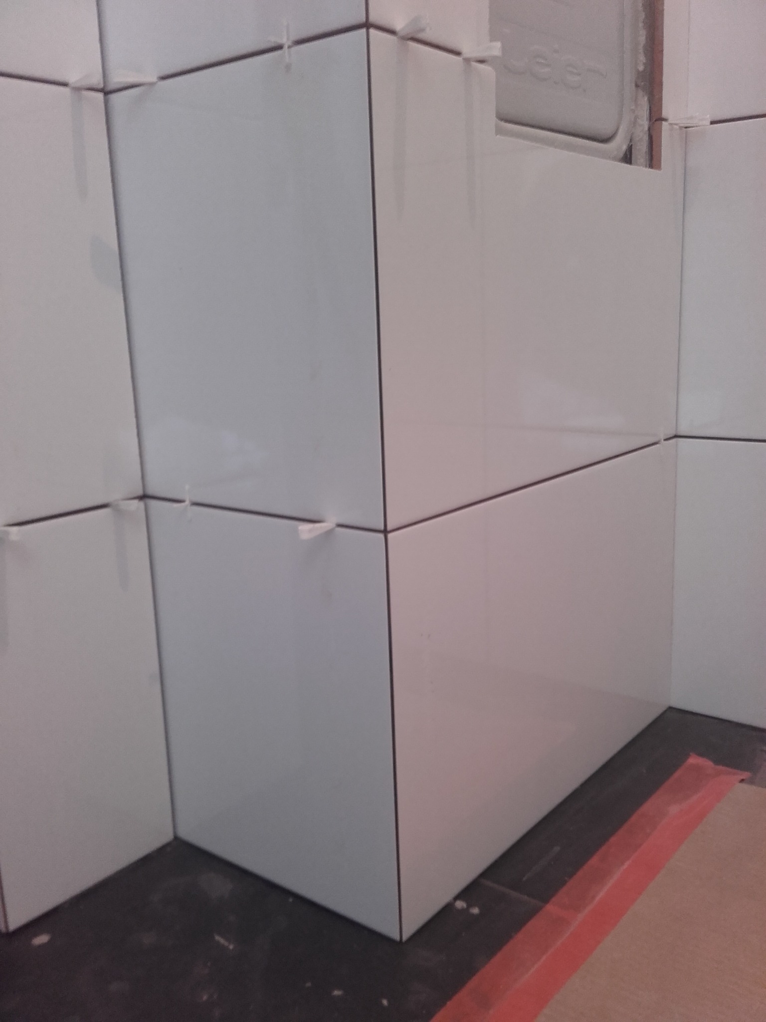 Świeżo położone, białe, prostokątne płytki ceramiczne na ścianie z użyciem plastikowych krzyżyków dystansowych, widoczne fragmenty podłogi zabezpieczonej folią.