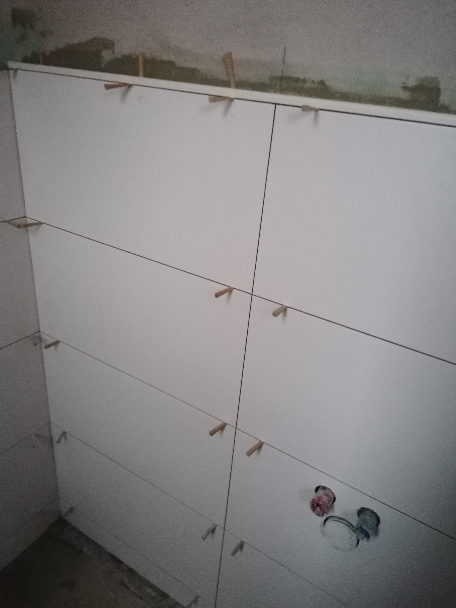 Świeżo położone, białe płytki na ścianie łazienki, z użyciem drewnianych krzyżyków dystansowych, widoczne otwory na armaturę, surowy stan wykończenia sufitu.