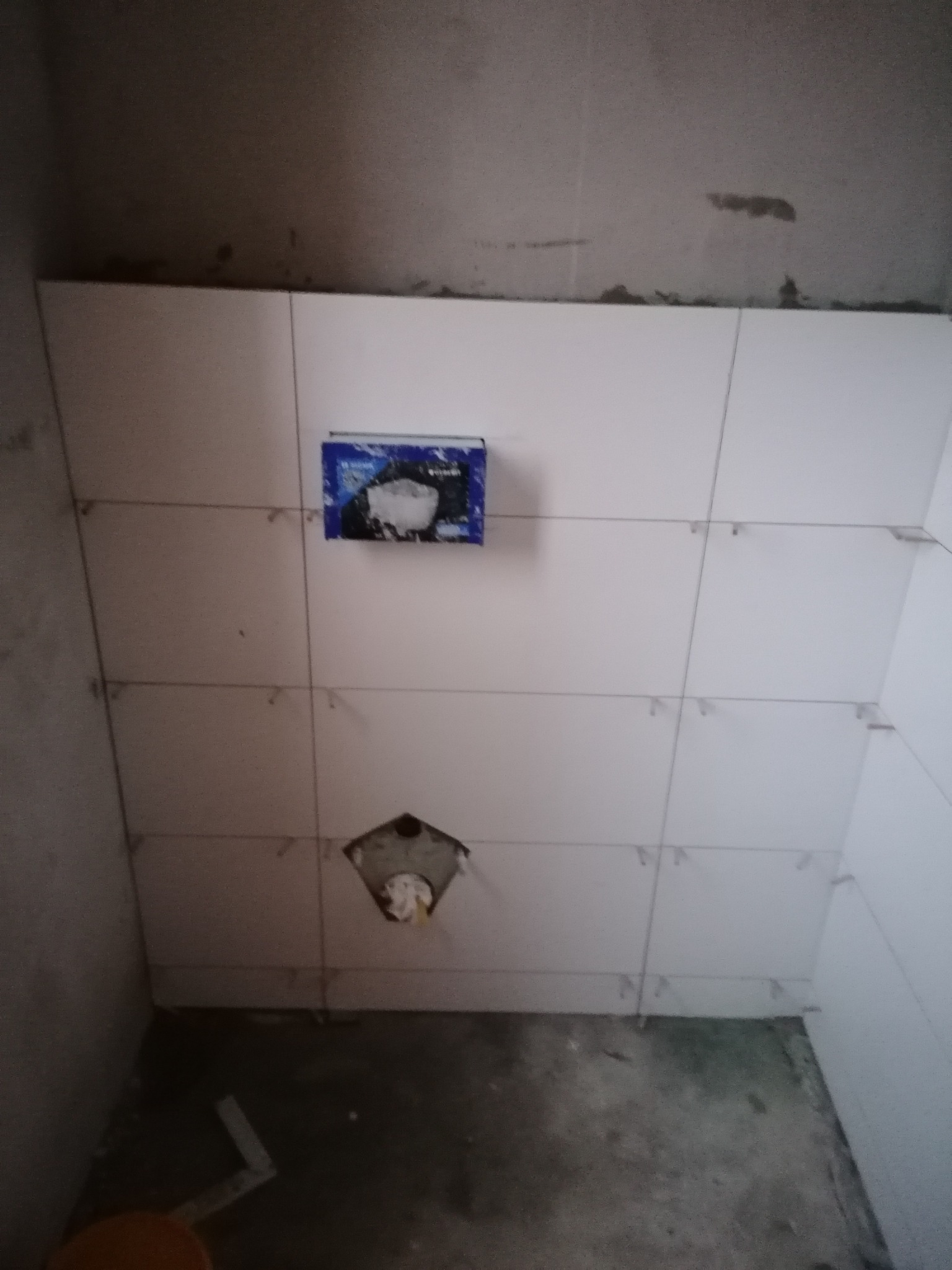 Ściana w trakcie układania jasnych płytek, widoczne krzyżyki dystansowe, puszka elektryczna i otwór na rury odpływowe, surowy stan podłogi.