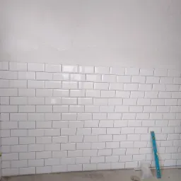 Ściana wyłożona białymi, prostokątnymi płytkami, z widoczną poziomicą przy ścianie, szpachlowana góra ściany, surowa podłoga i resztki materiałów budowlanych.