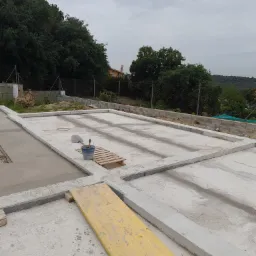 Wylany fundament z betonowymi obrzeżami, widoczny fragment żółtej deski i wiadro z narzędziami na palecie, w tle drzewa i pochmurne niebo.