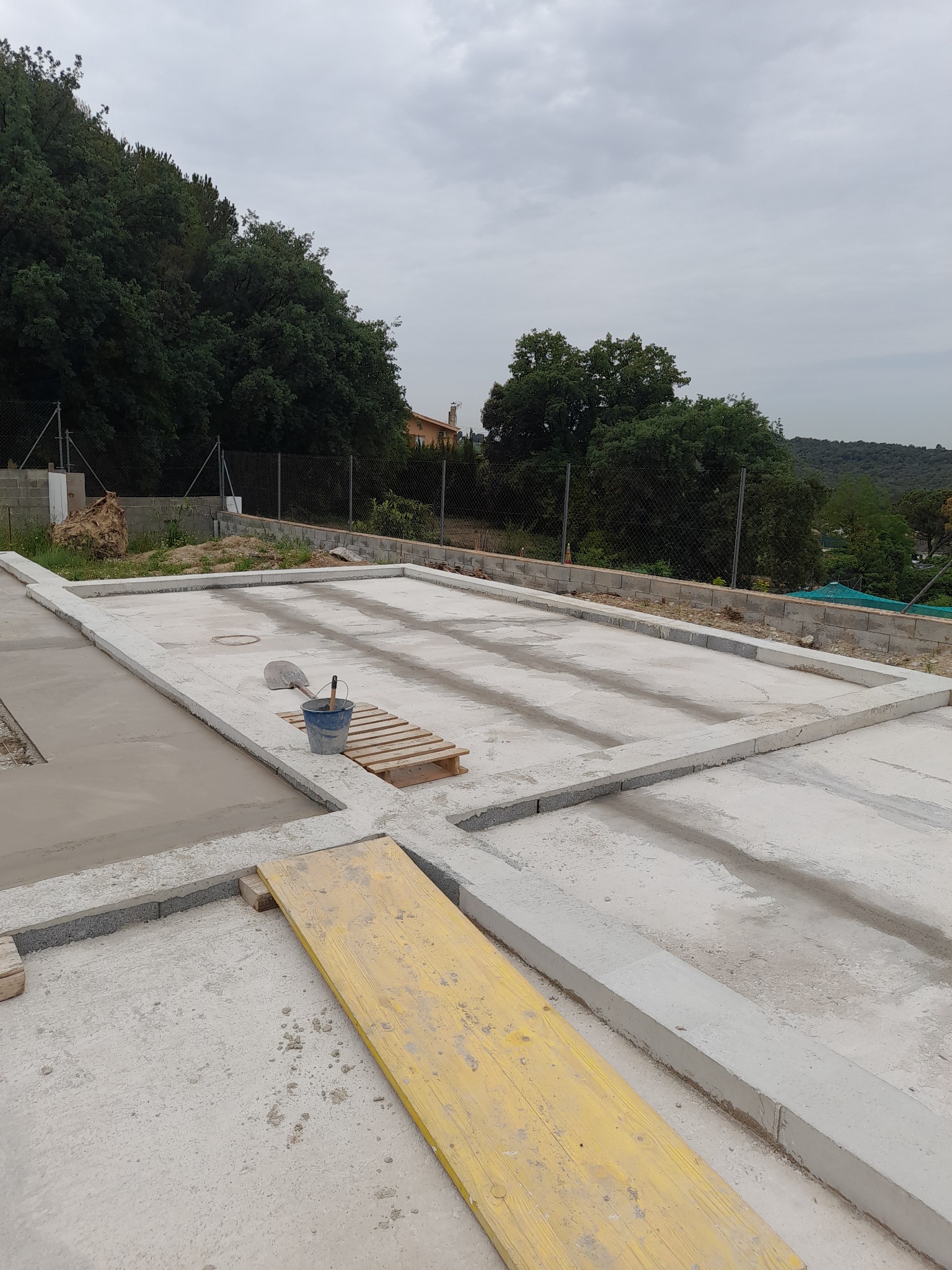 Wylany fundament z betonowymi obrzeżami, widoczny fragment żółtej deski i wiadro z narzędziami na palecie, w tle drzewa i pochmurne niebo.