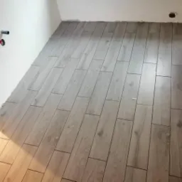 Ułożona podłoga z płytek imitujących drewno w jasnym odcieniu, widoczne wystające rury na ścianie i otwór w ścianie przy podłodze.