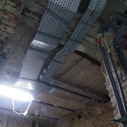 RefNeck Sp. z o.o. - Sufit w zabytkowej kamienicy z widocznymi metalowymi korytkami kablowymi ułożonymi równolegle, białymi przewodami elektrycznymi oraz rurami instalacyjnymi w szarym kolorze.