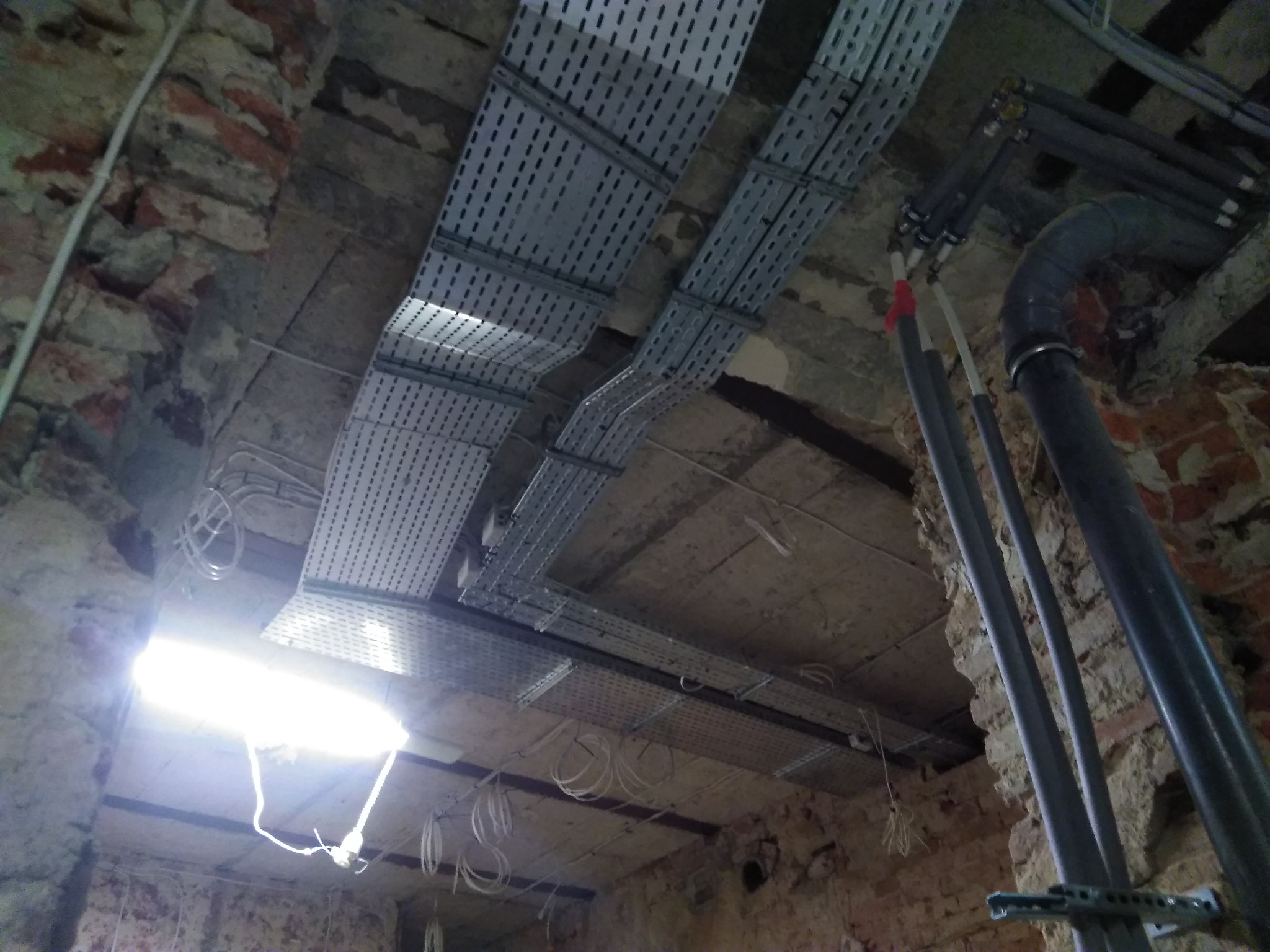Sufit w zabytkowej kamienicy z widocznymi metalowymi korytkami kablowymi ułożonymi równolegle, białymi przewodami elektrycznymi oraz rurami instalacyjnymi w szarym kolorze.
