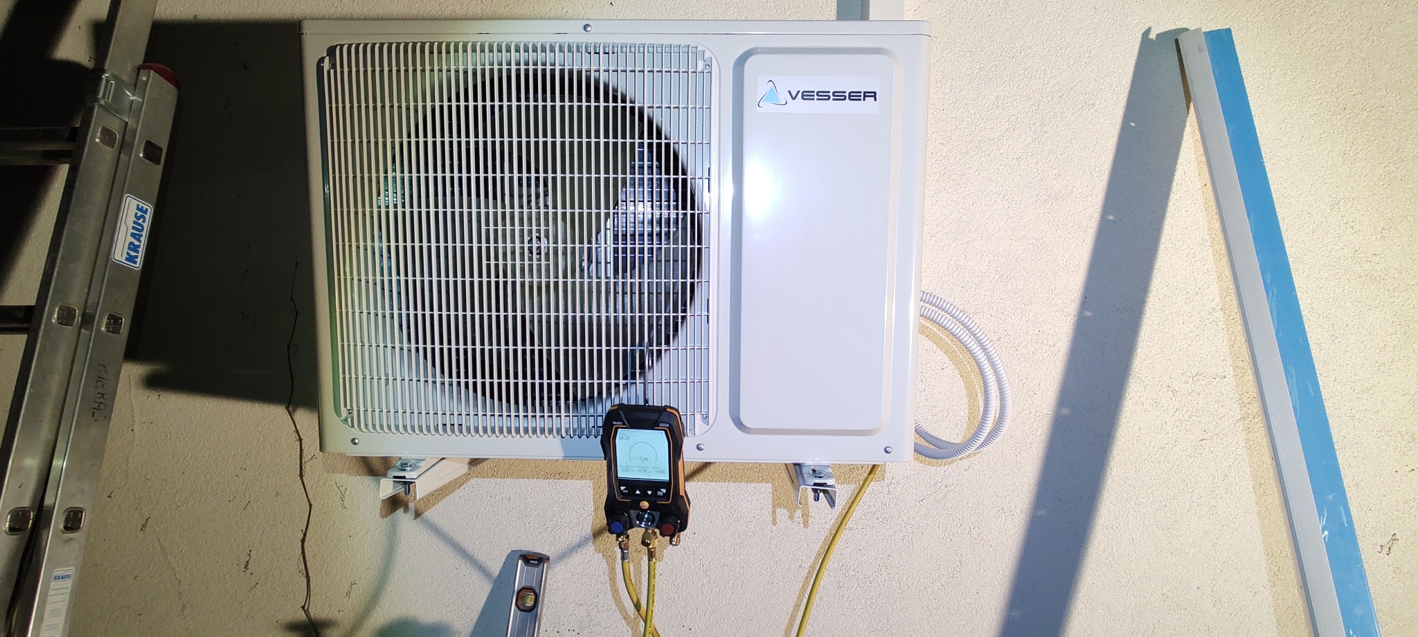 Zewnętrzna jednostka klimatyzacji marki Vesser zamontowana na ścianie budynku, podłączona do manometru cyfrowego podczas przeglądu technicznego, widoczna poziomica i drabina Krause