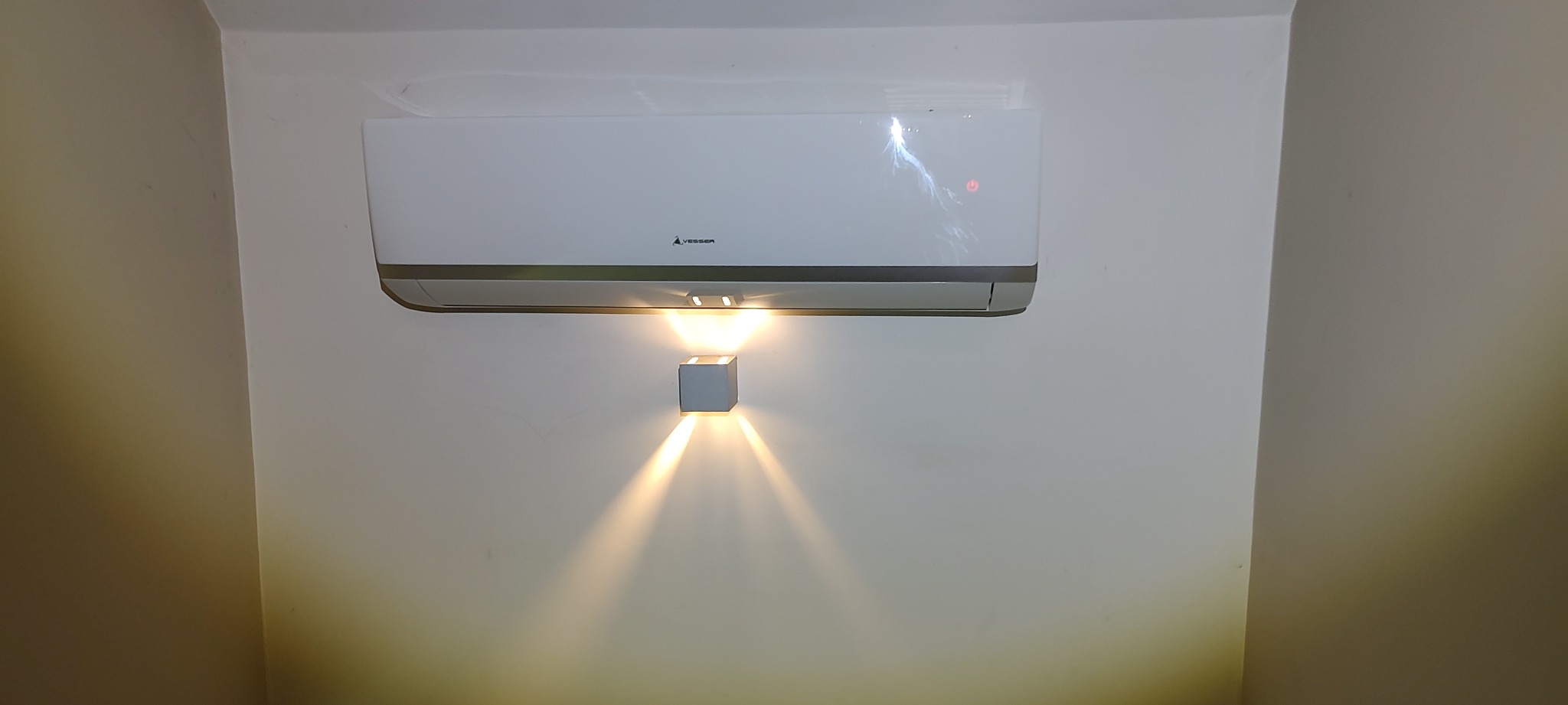 Biały klimatyzator marki Vesser zamontowany na ścianie nad kwadratową lampą ścienną, oświetlającą ścianę ciepłym światłem.