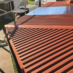 Czerwony blaszany dach z widocznymi łączeniami i fragmentem orynnowania, na tle rusztowania i budynków.