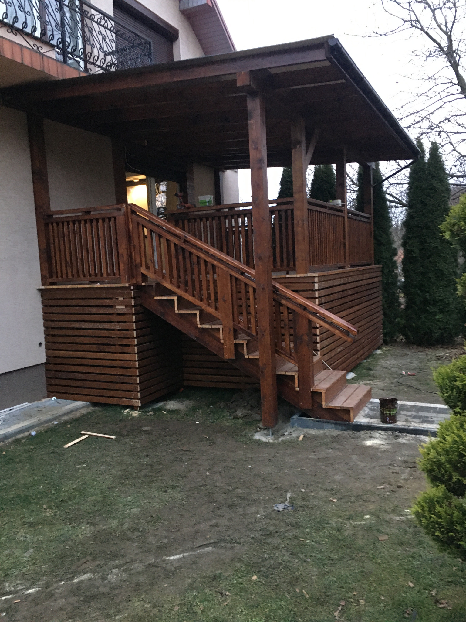 Drewniany taras z zadaszeniem i schodami, wykończony w ciemnym brązie, przylegający do budynku z balkonem z kutą balustradą, widok z poziomu gruntu na trawnik z resztkami materiałów budowlanych.