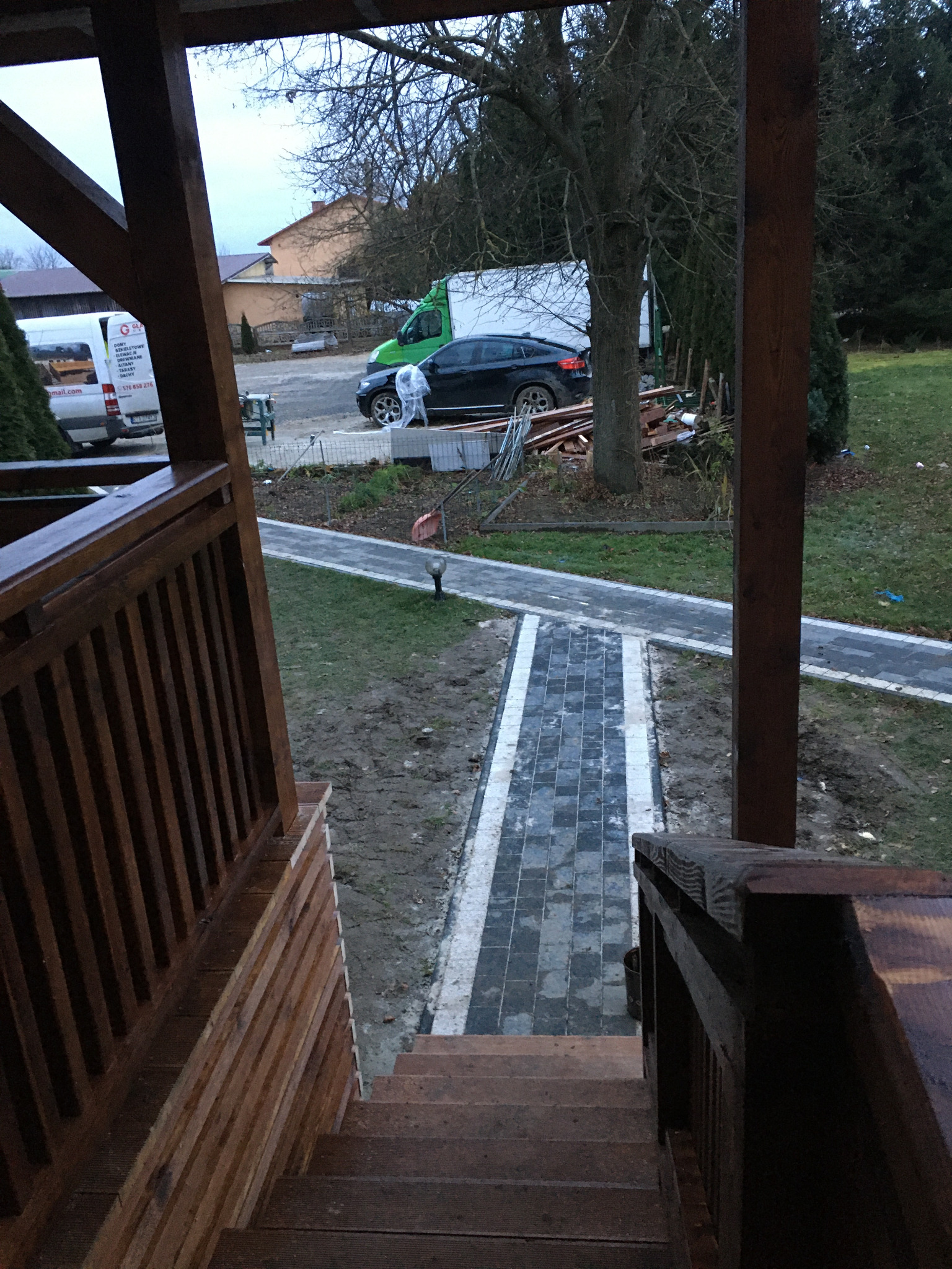 Widok z drewnianego tarasu na ścieżkę z kostki brukowej prowadzącą do posesji, z widocznymi samochodami i stertą drewna w tle.