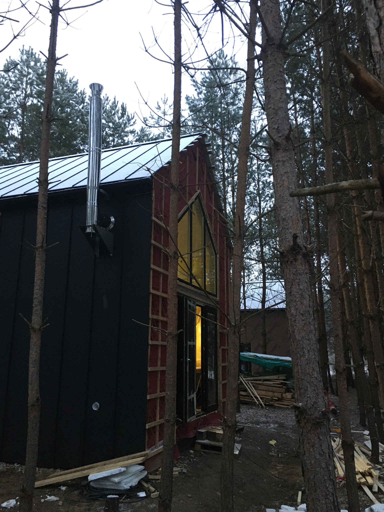 Niedokończony budynek z czarną elewacją i kominem stalowym, widoczny przez las, z oświetlonym wnętrzem i elementami konstrukcyjnymi.