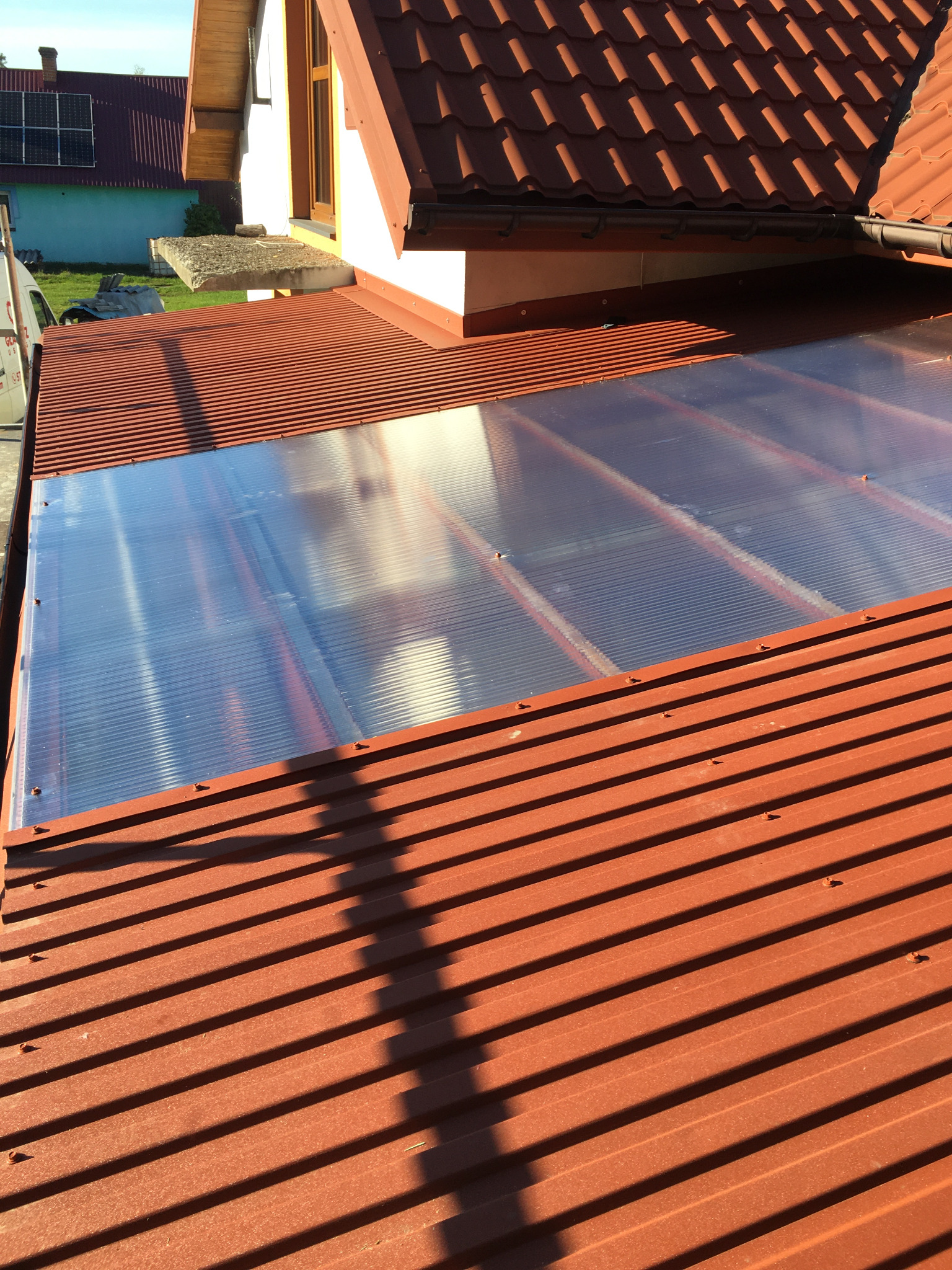 Czerwony dach z blachy trapezowej z przezroczystym świetlikiem z poliwęglanu, widoczne mocowania, w tle fragment domu z panelami słonecznymi.