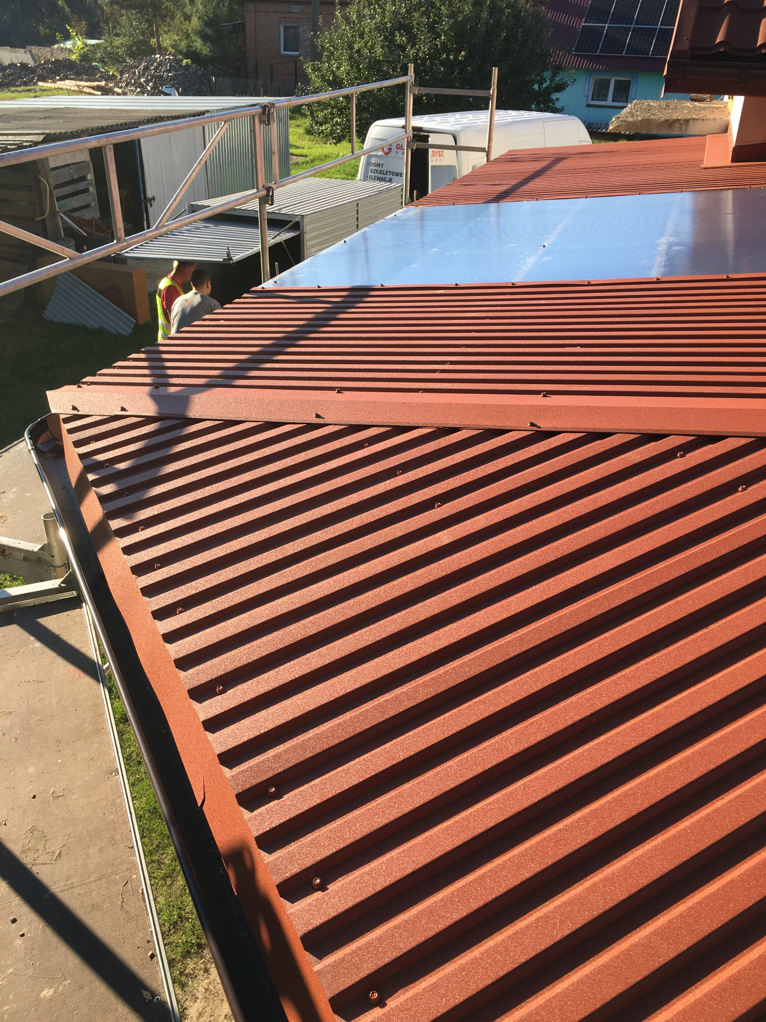 Czerwony blaszany dach z widocznymi łączeniami i fragmentem orynnowania, na tle rusztowania i budynków.