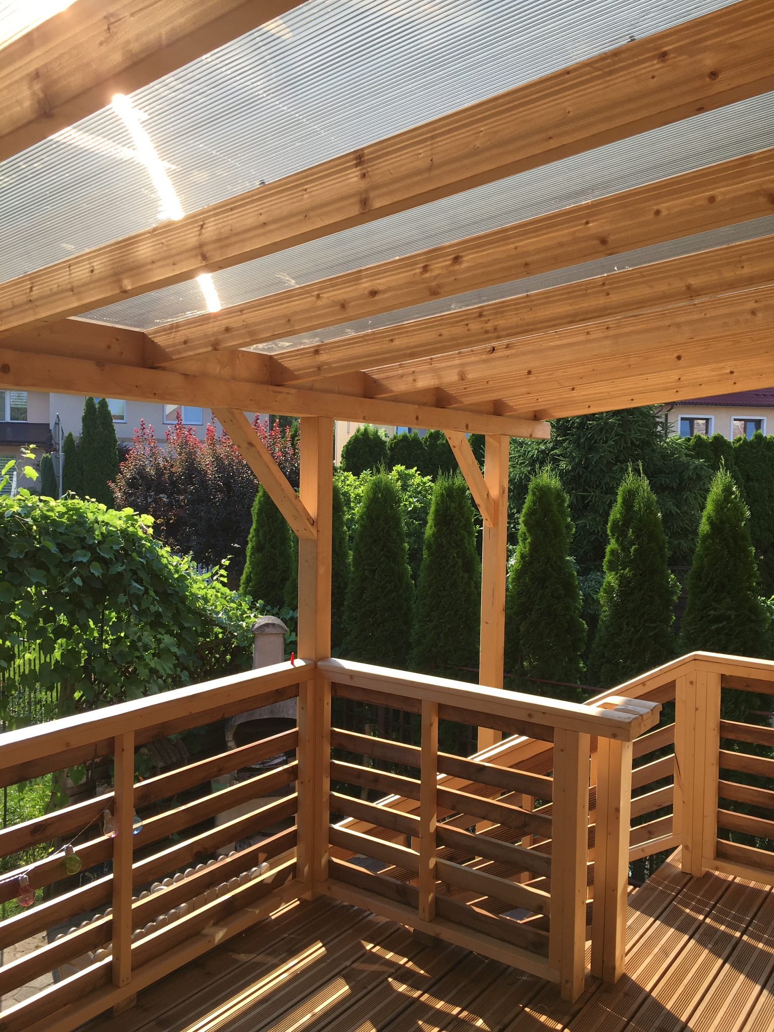 Drewniany taras z zadaszeniem z przezroczystego tworzywa, widok z wnętrza, w tle zieleń drzew i krzewów.