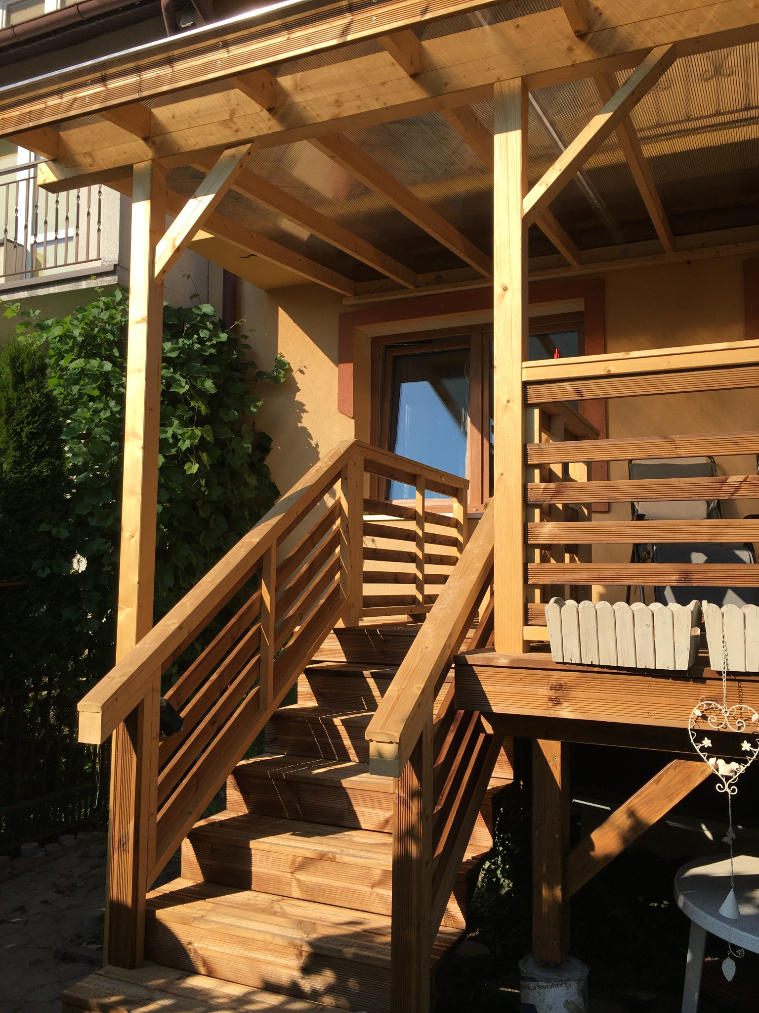 Drewniany taras z zadaszeniem z przezroczystego tworzywa, schody z balustradą, donica z kwiatami i ozdobny wisiorek w kształcie serca.