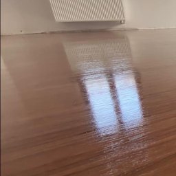 Grindelwald - Błyszcząca, świeżo polakierowana podłoga drewniana w pokoju z oknem i grzejnikiem, widoczne odbicie okna w lakierze.