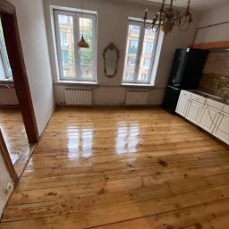 Grindelwald - Wycyklinowana podłoga z desek w pokoju z dwoma oknami, widoczne ślady po poprzednich zabrudzeniach, sufit ze skosem i wiszącymi lampami.